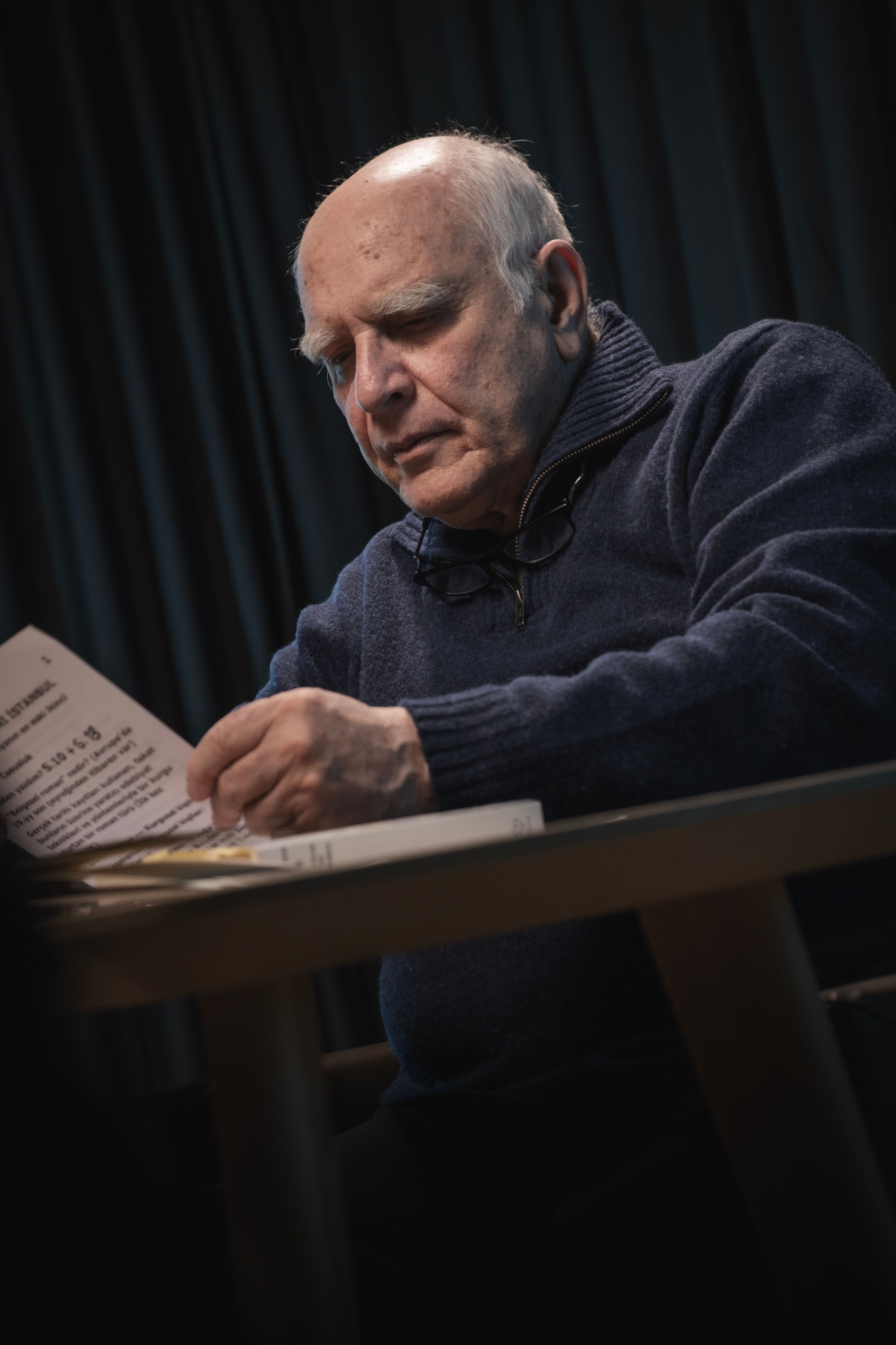 An elderly man in a sweater sits at a table reading a document indoors.
