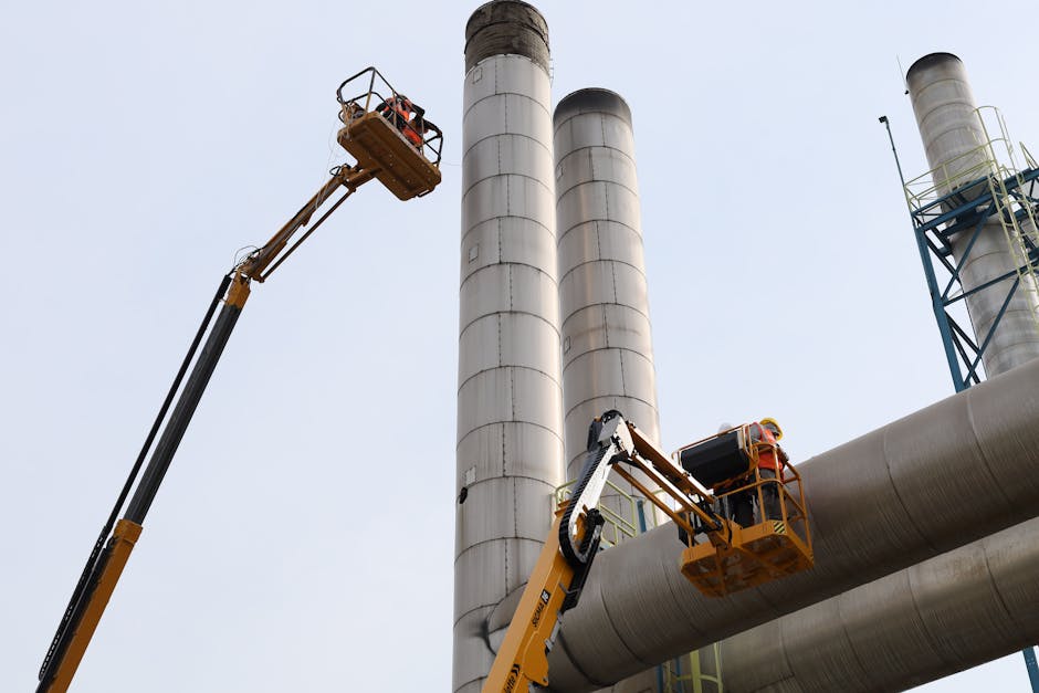 Workers using lifts for maintenance near towering factory chimneys against a clear sky.