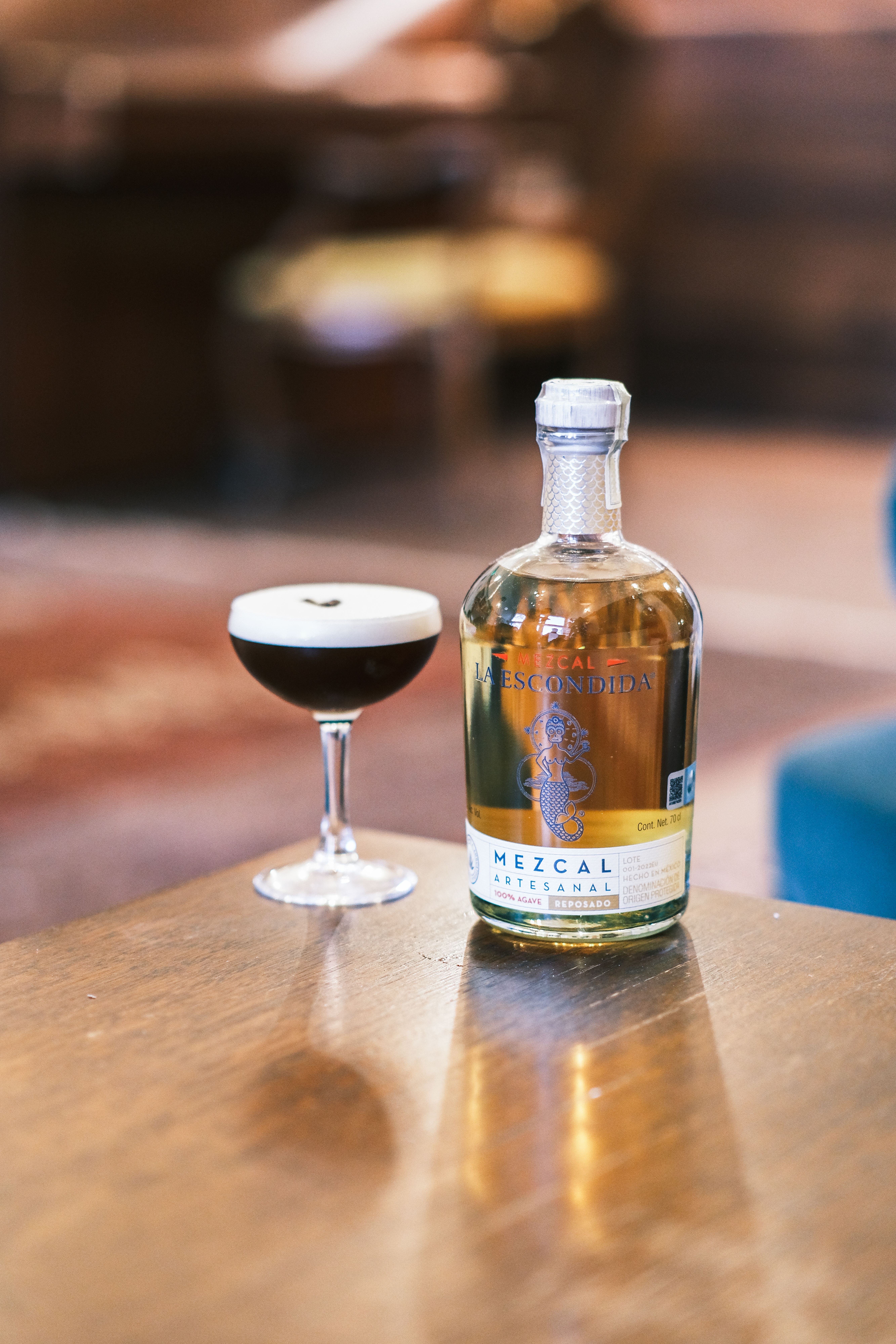A bottle of artisan mezcal beside a cocktail on a wooden table, emphasizing craft beverage culture.