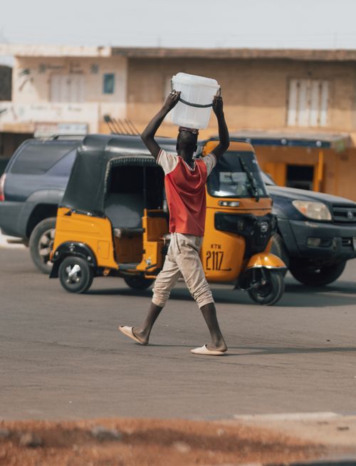 Free A man carries a water bucket on his head while crossing a busy urban street. Stock Photo