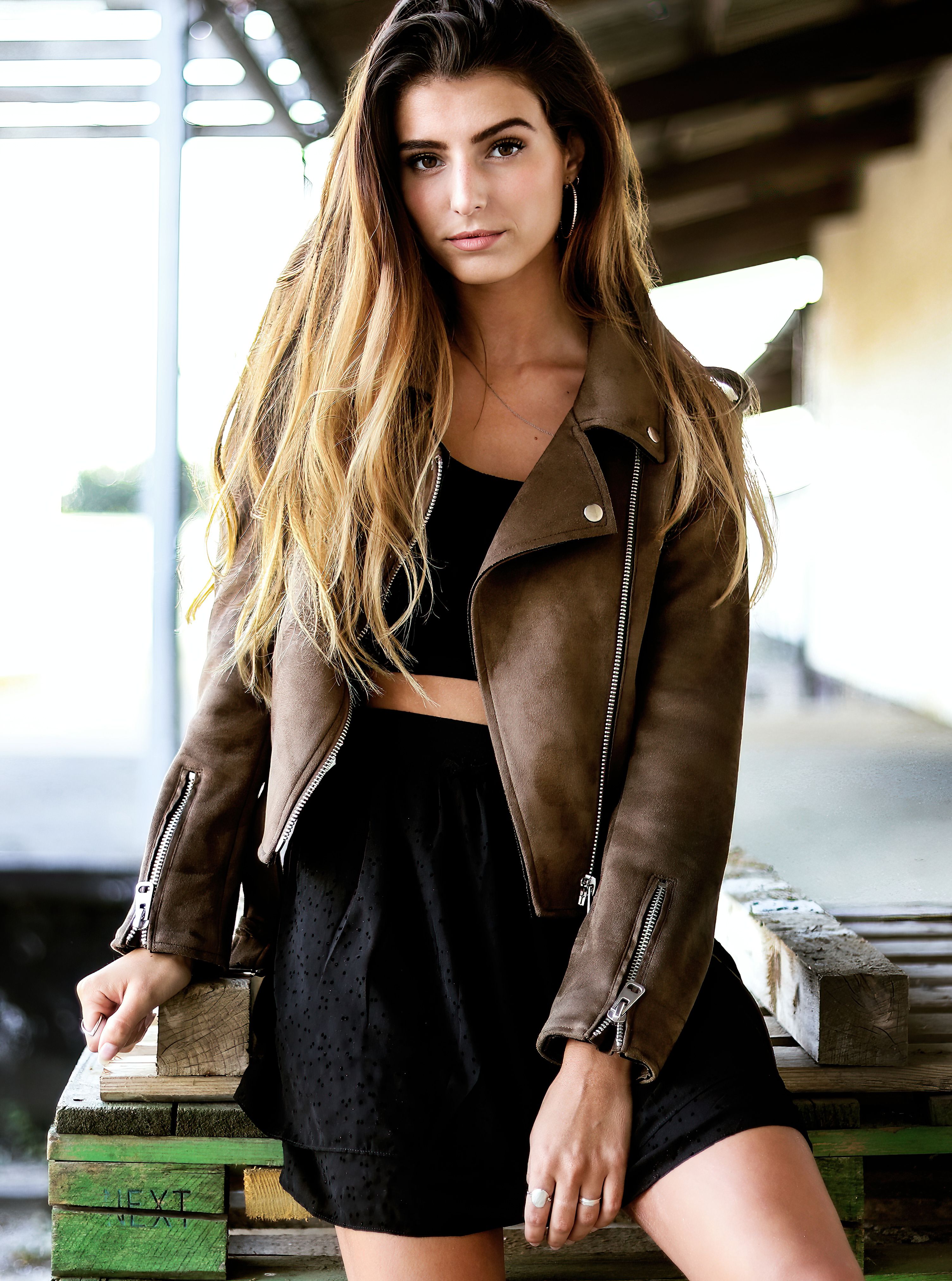 Young woman with long blonde hair wearing a leather jacket poses confidently outdoors.