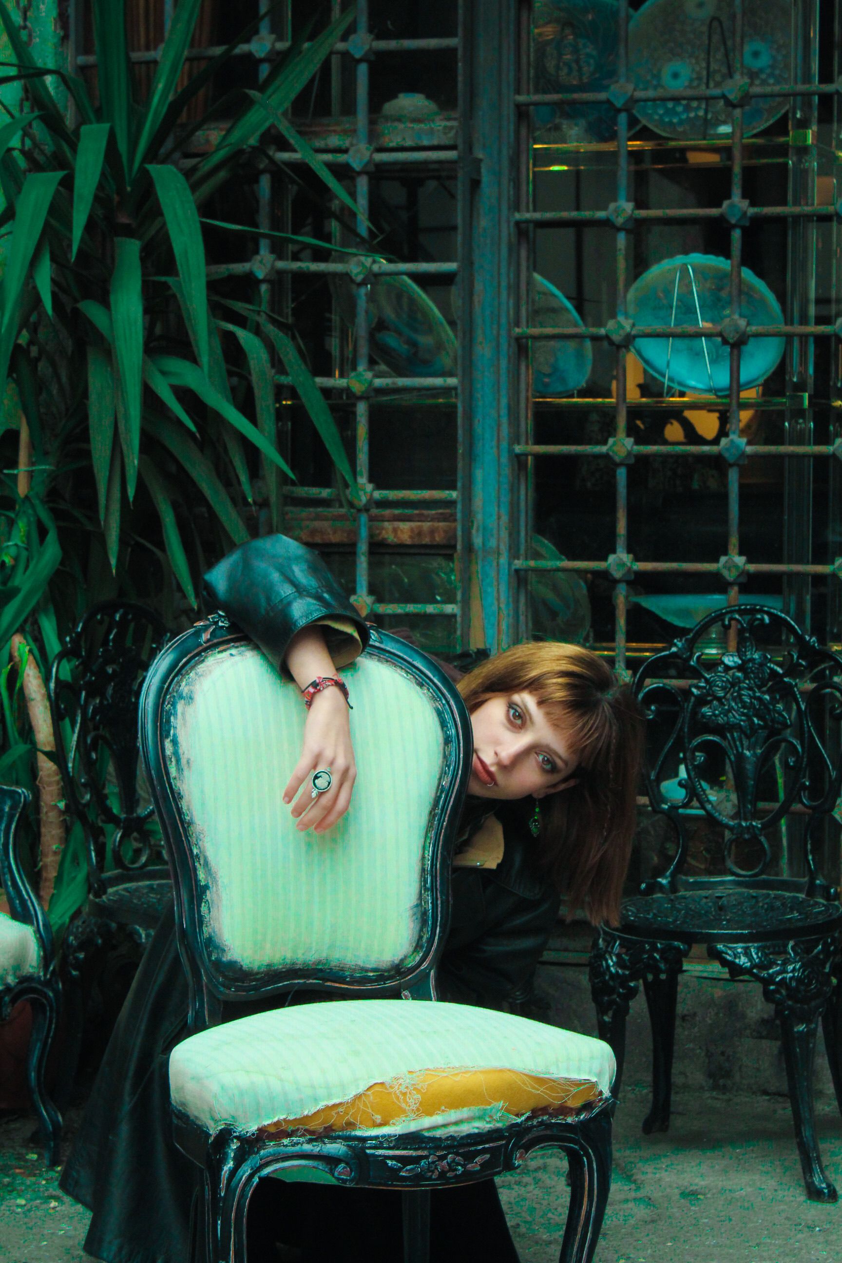 Woman posing with vintage chair in an outdoor setting with gothic metal elements and greenery.