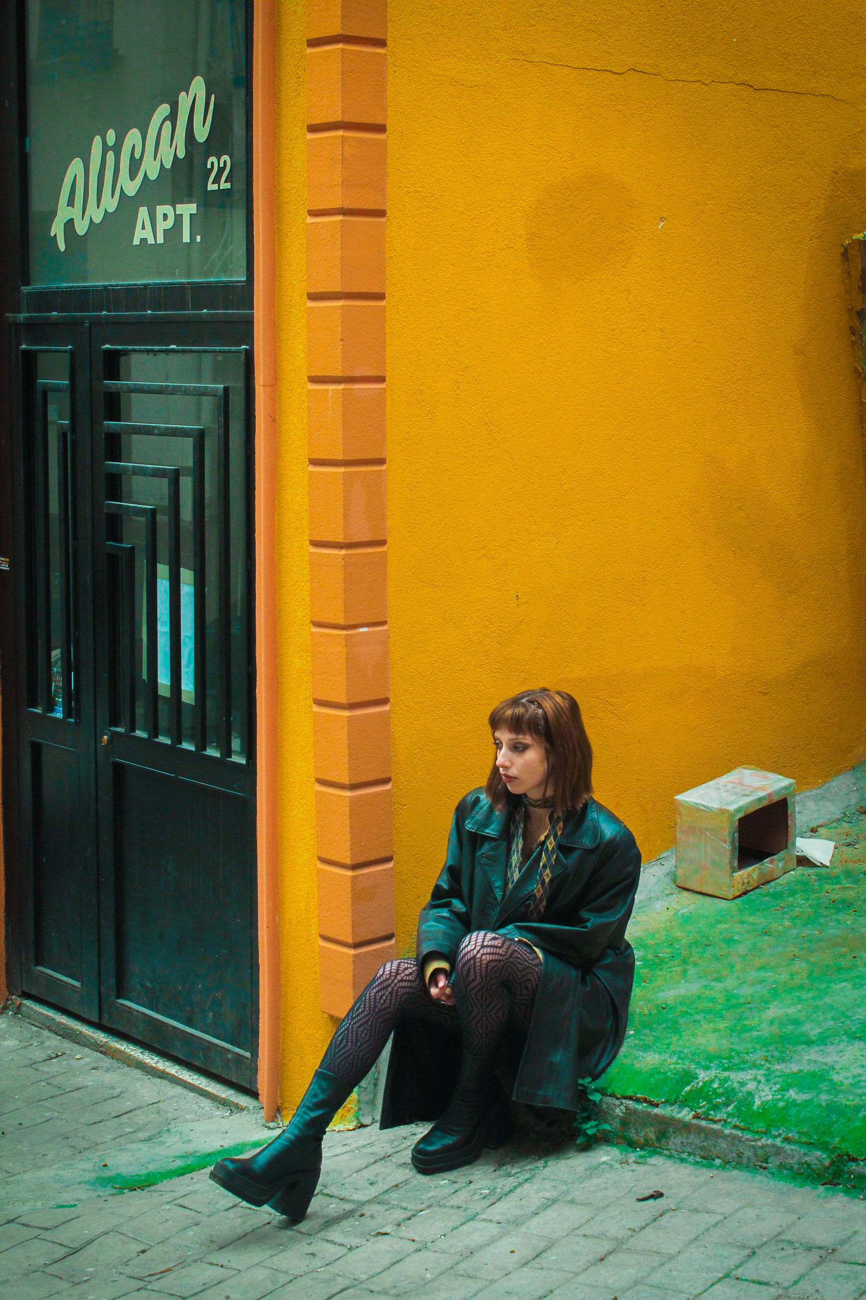 Woman in green leather jacket sitting by a bright yellow wall in urban setting.