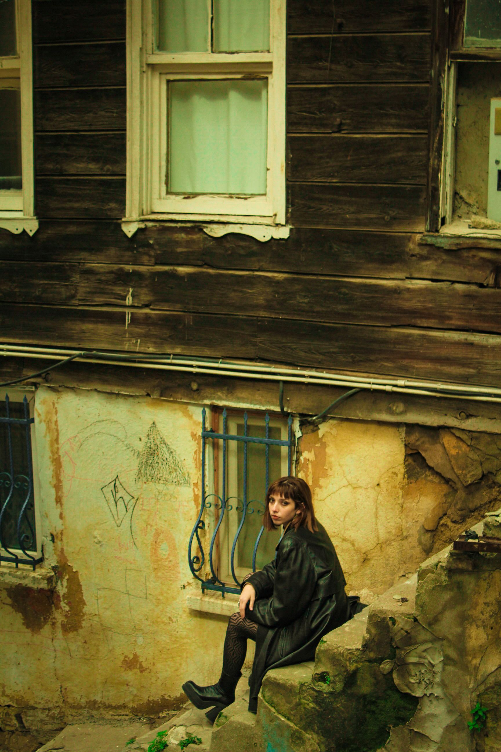 A moody portrait of a woman sitting beside an abandoned, decaying building.