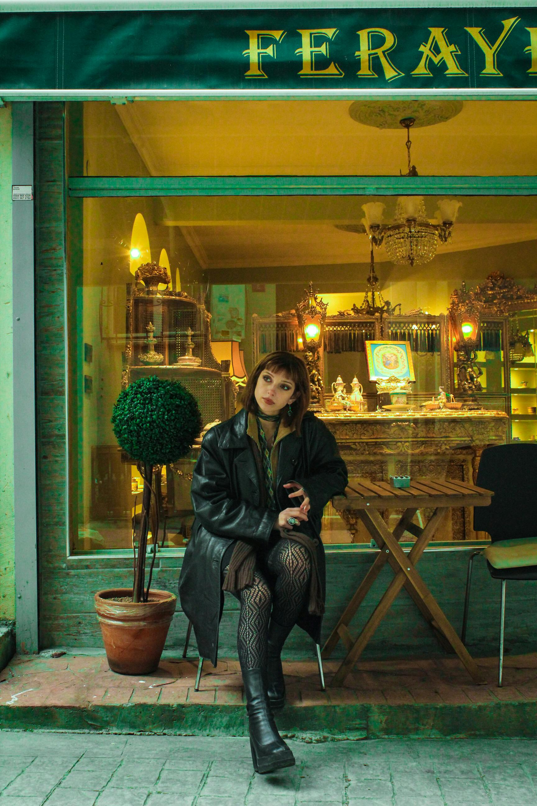 A woman sits in front of an antique shop, creating a cozy urban scene with warm tones.