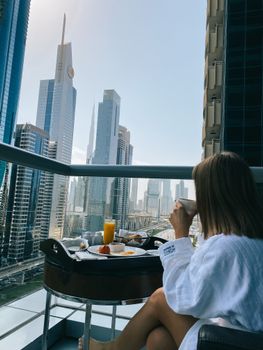 Woman in bathrobe enjoying breakfast on a balcony with Dubai skyline view.