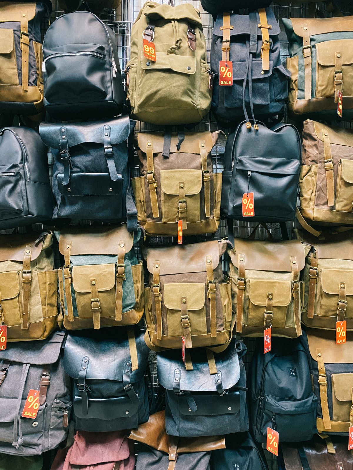 A wall of canvas and leather backpacks with buckled flaps hanging in a shop, some with sale tags