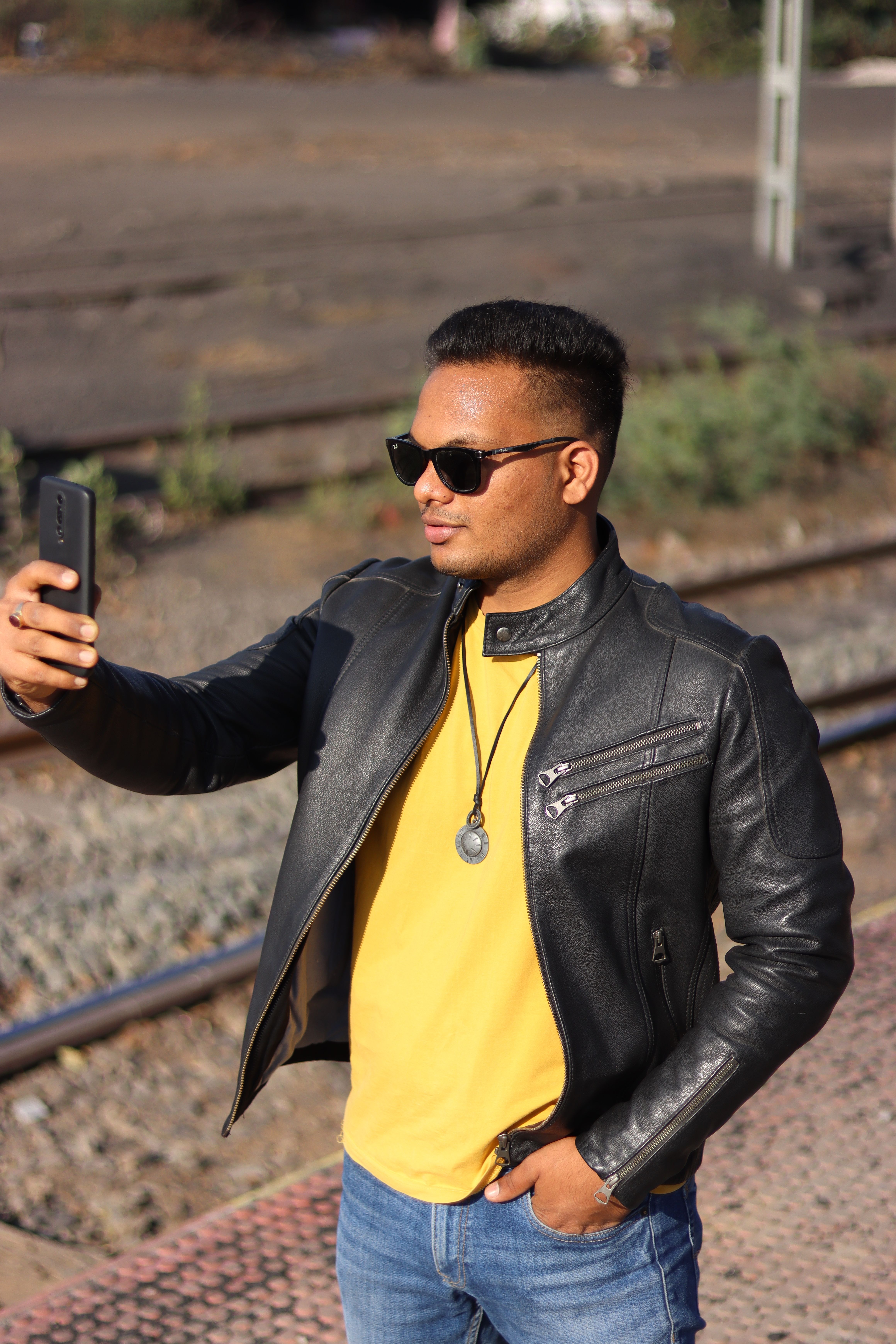 Stylish man in sunglasses taking a selfie with a smartphone while wearing a leather jacket outdoors.