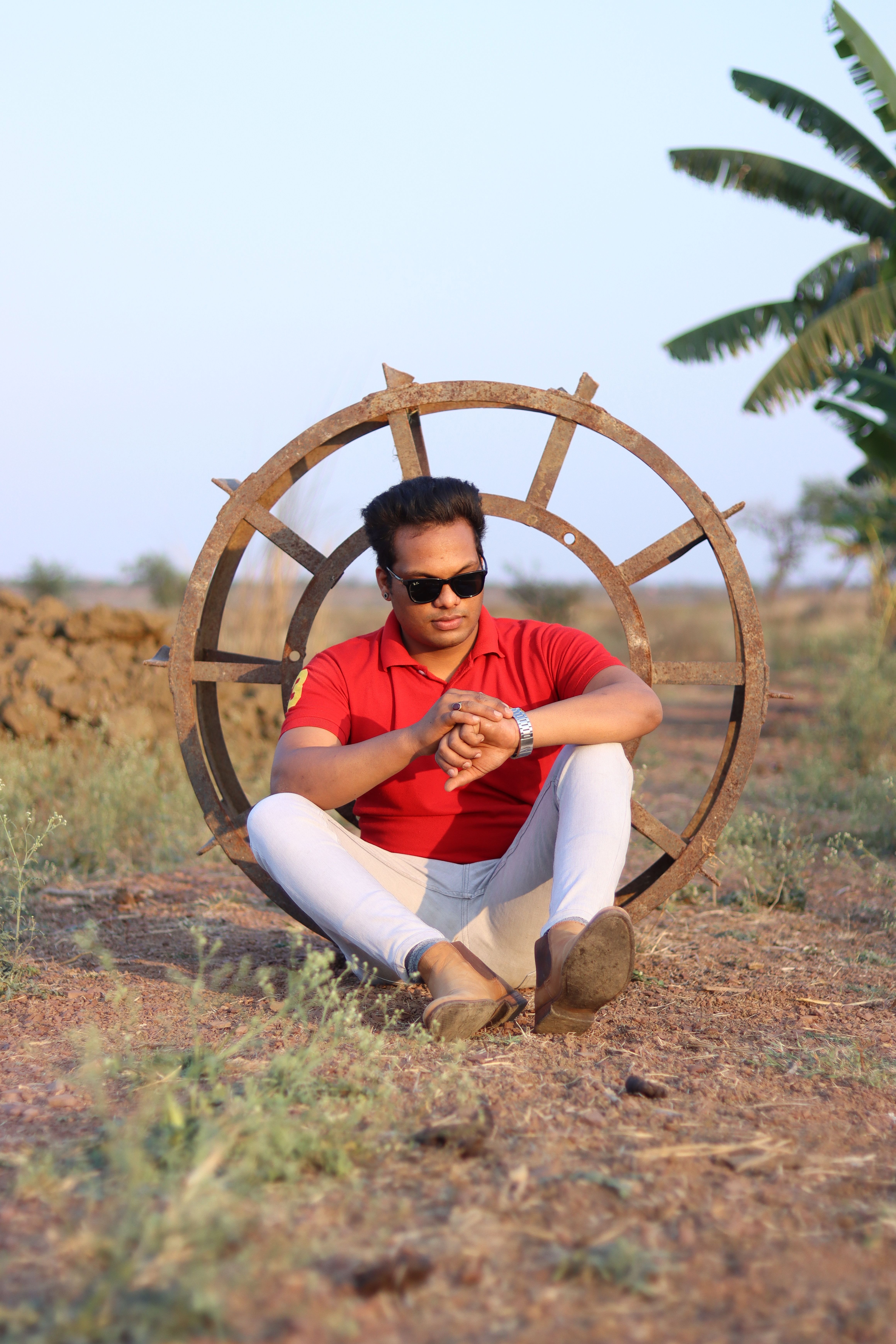Portrait of a fashionable man sitting against a rustic wheel in nature, exuding style.