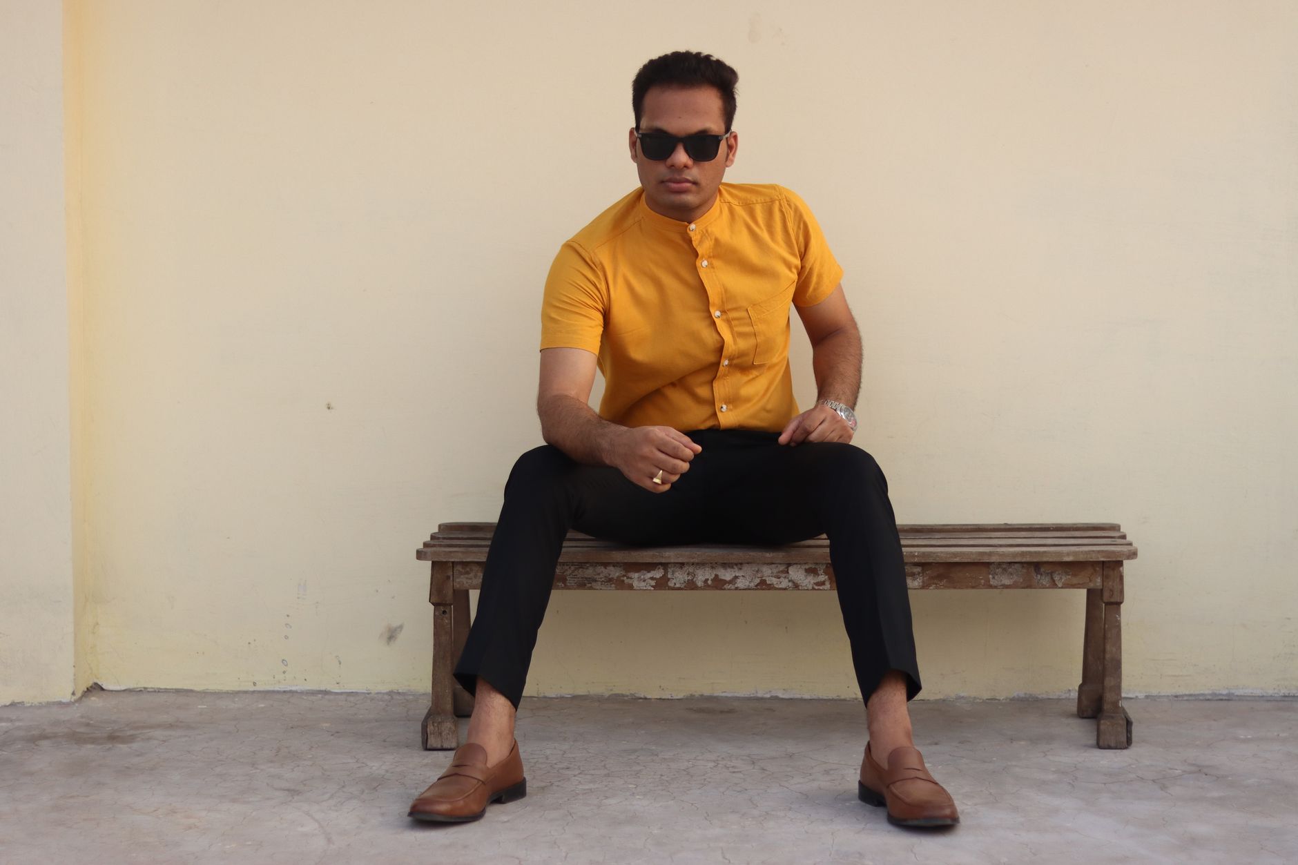 Fashionable man in yellow shirt and sunglasses sitting on bench outdoors.