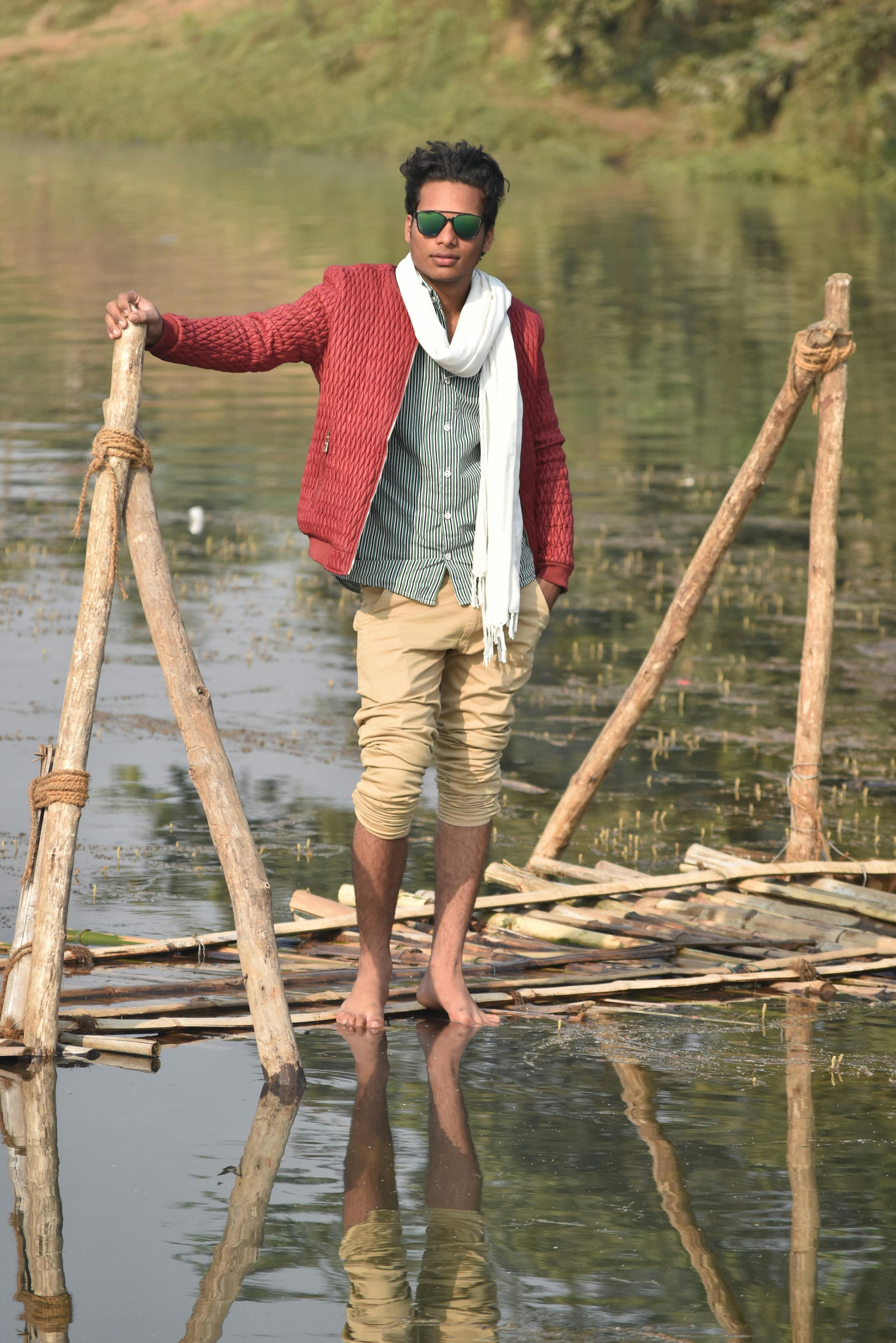Young man in fashionable attire poses confidently on a rustic bridge over a serene river.