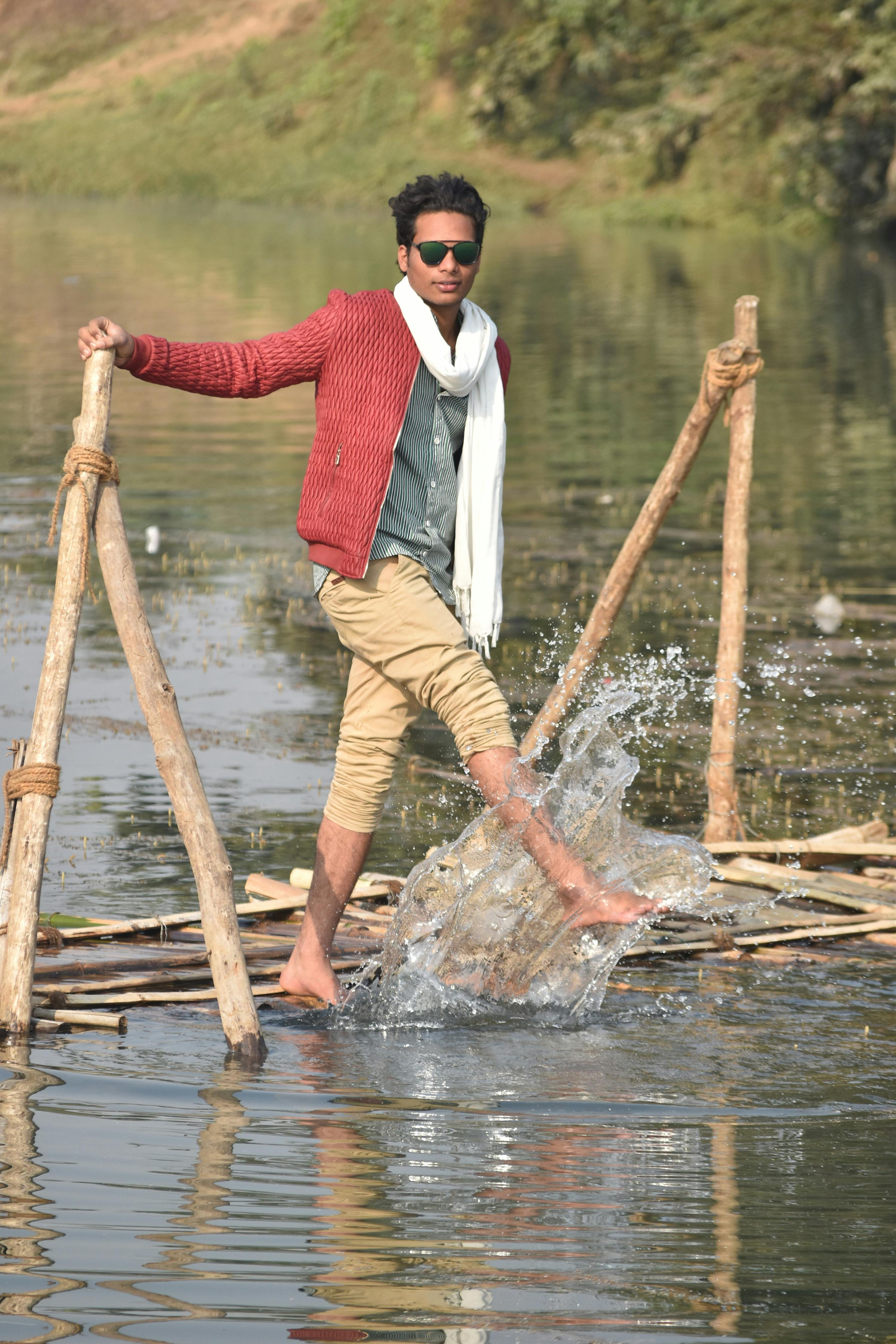 Man in stylish attire kicks water on a rustic jetty, showcasing fashion and action in an outdoor scene.