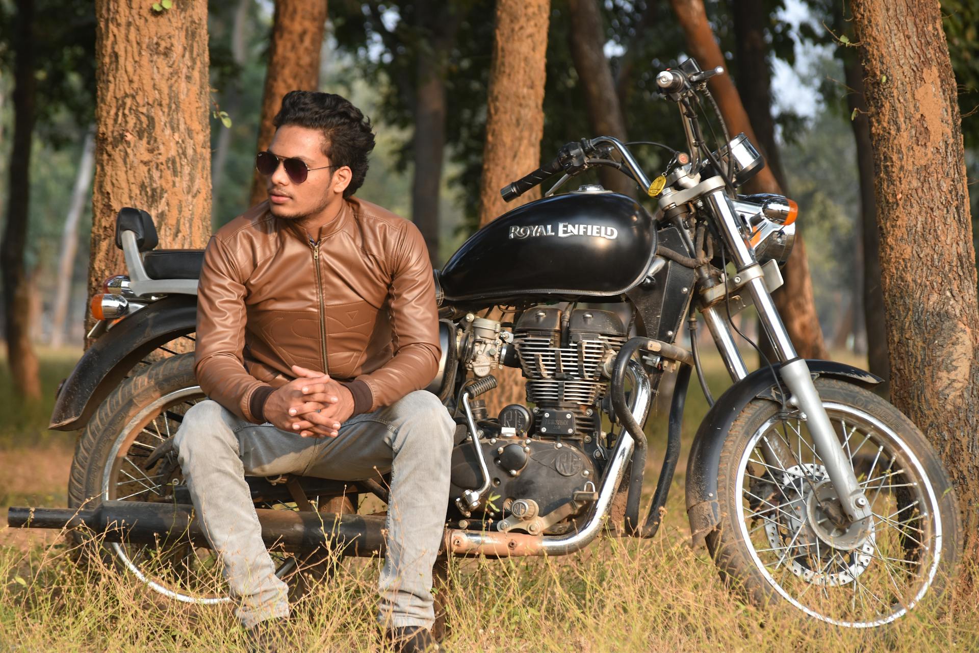 Casual man in leather jacket sitting on a Royal Enfield motorcycle in a sunny forest setting.