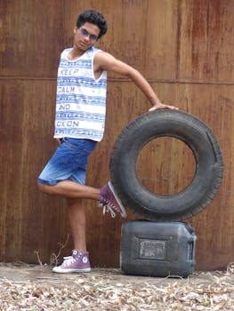 Young man in casual attire posing with a tire against a rustic background outdoors.