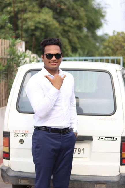 Smiling man in white shirt and sunglasses making shaka gesture beside a white car.
