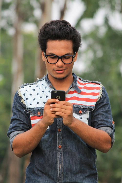 Free Young man wearing denim shirt with stars and stripes, using smartphone outdoors. Stock Photo