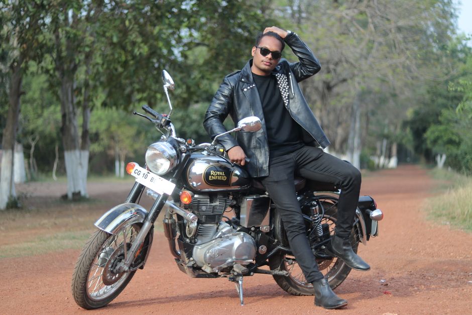 Stylish man in black leather jacket posing on Royal Enfield motorcycle outdoors.