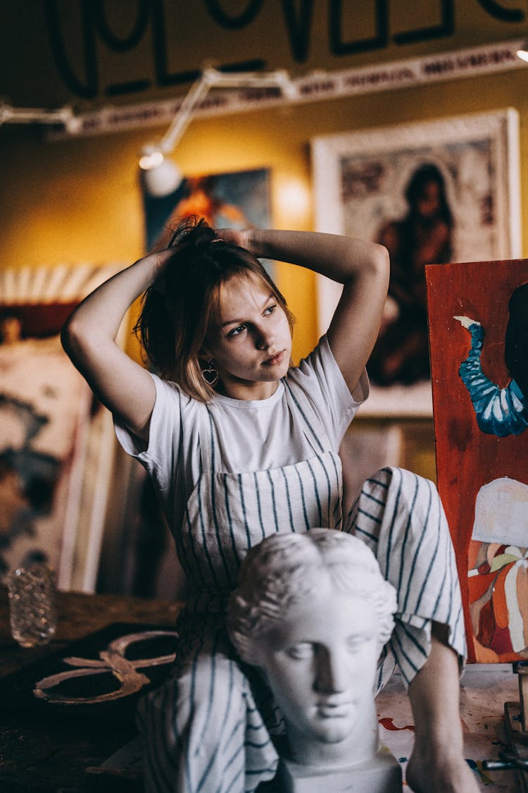 Blond Girl Wearing Striped Dungarees Posing In An Artists Studio With Pictures And A Sculpture