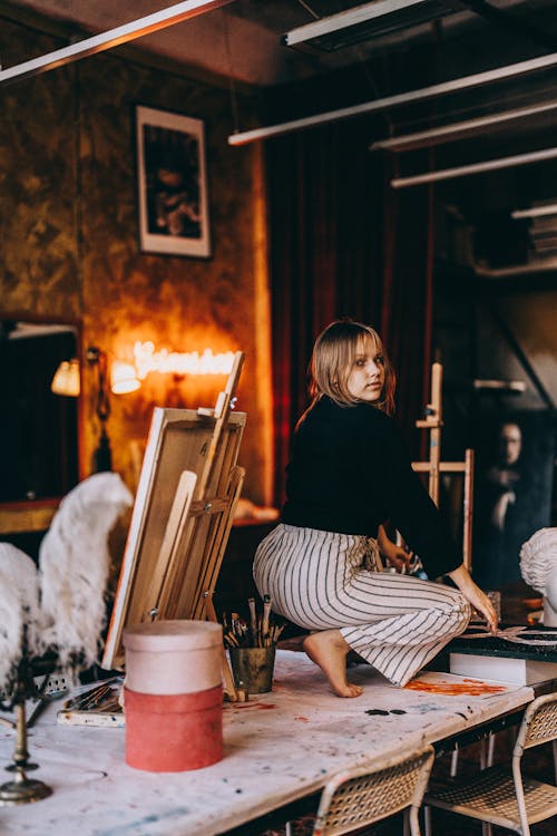 Girl Wearing Striped Trousers Crouching Barefoot on a Table in an Artists Studio