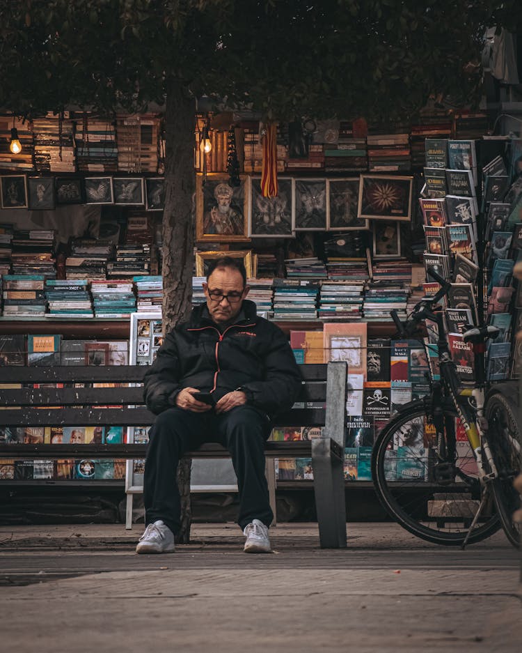 Looking At Smartphone Man Sit On Bench By Stall With Books