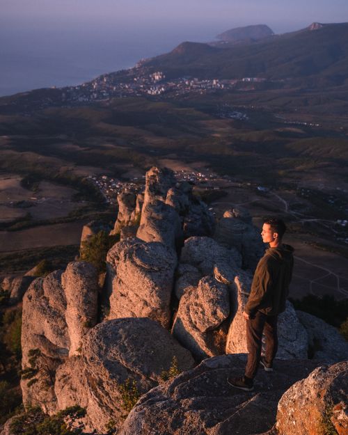 Free Man standing on rocky cliff overlooking expansive landscape and town at sunset, embracing adventure. Stock Photo