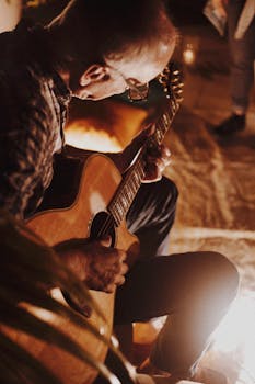 Man playing acoustic guitar by firelight creating a cozy atmosphere.