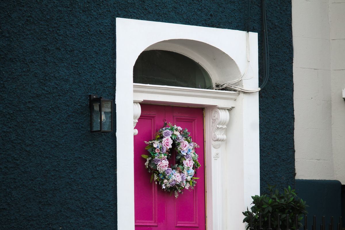 A vibrant pink door adorned with a floral wreath, set against a textured blue wall, exudes charm and elegance. Stock Photo.