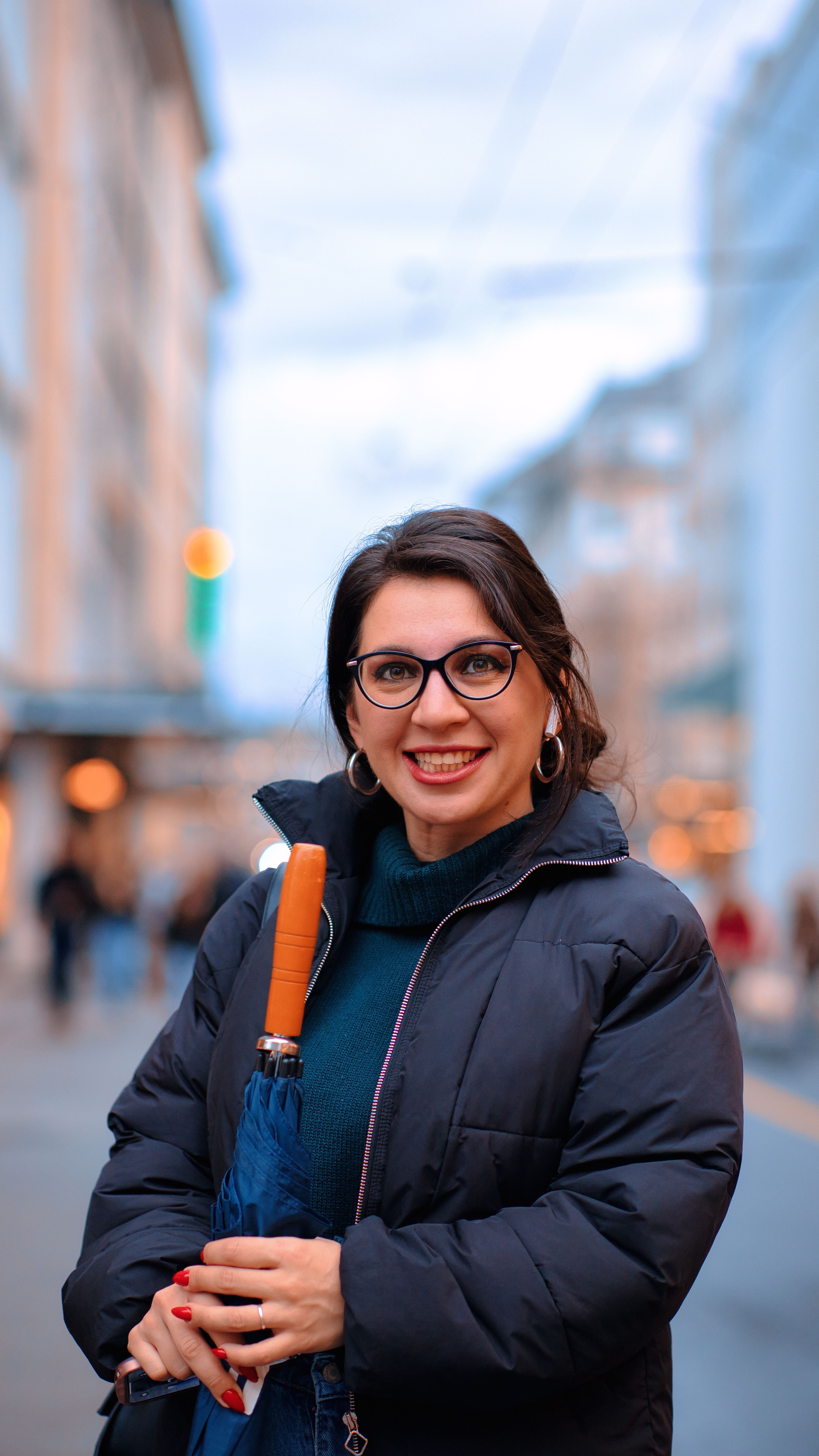 A cheerful woman in warm clothing holds an umbrella on a vibrant city street.