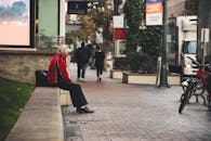 一位老人穿着红色夹克，独自坐在城市街道的长凳上，捕捉着城市的孤独