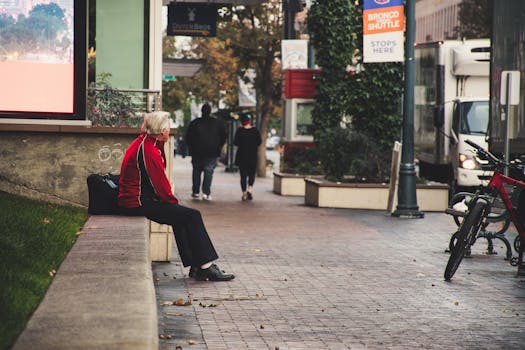 一位老人穿着红色夹克，独自坐在城市街道的长凳上，捕捉着城市的孤独