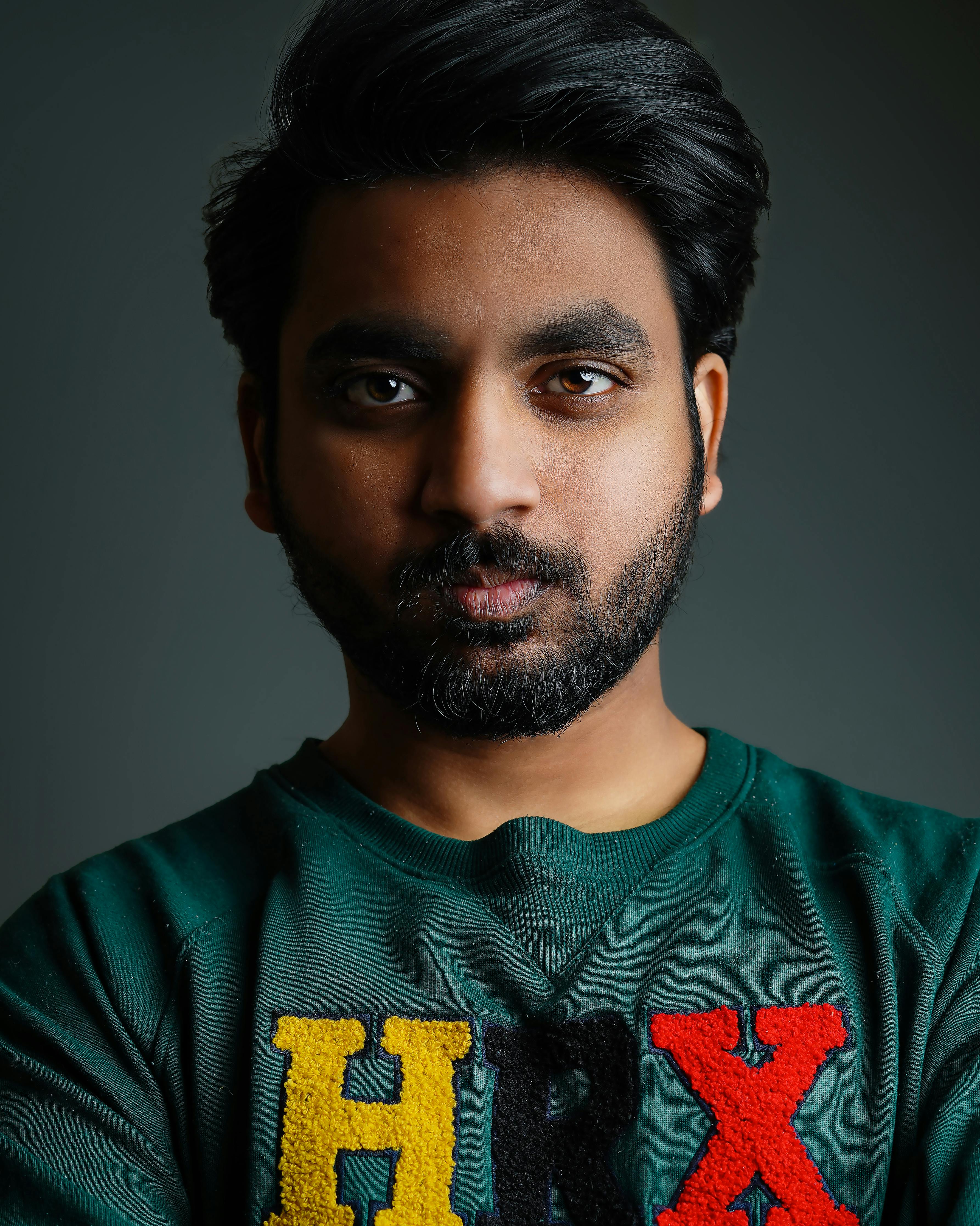 Close-up headshot of a South Asian man with stubble and black hair in a colorful sweatshirt on a green background.