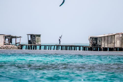 Free Kitesurfer gliding along a rustic pier, surrounded by vibrant blue ocean waters under a clear sky. Stock Photo