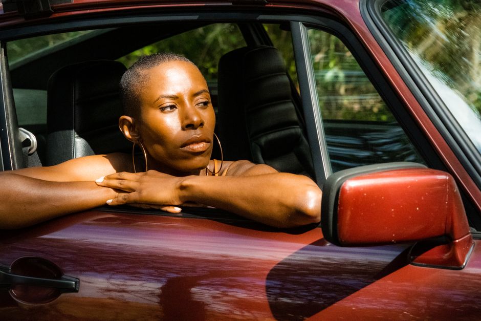 A woman with short hair gazes thoughtfully out the window of a parked car, bathed in sunlight.