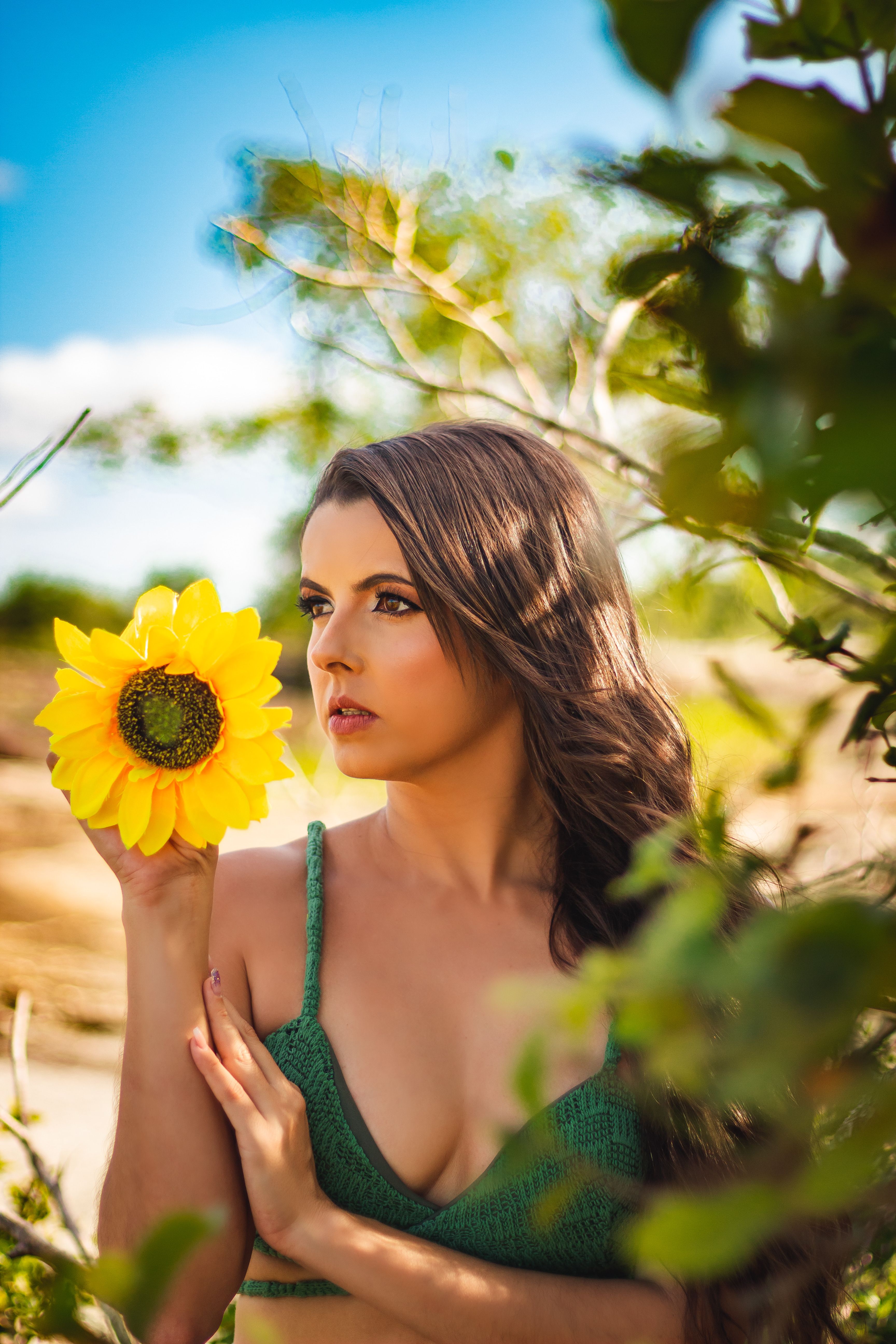 A young woman in a green top holds a vibrant sunflower outdoors on a sunny summer day.