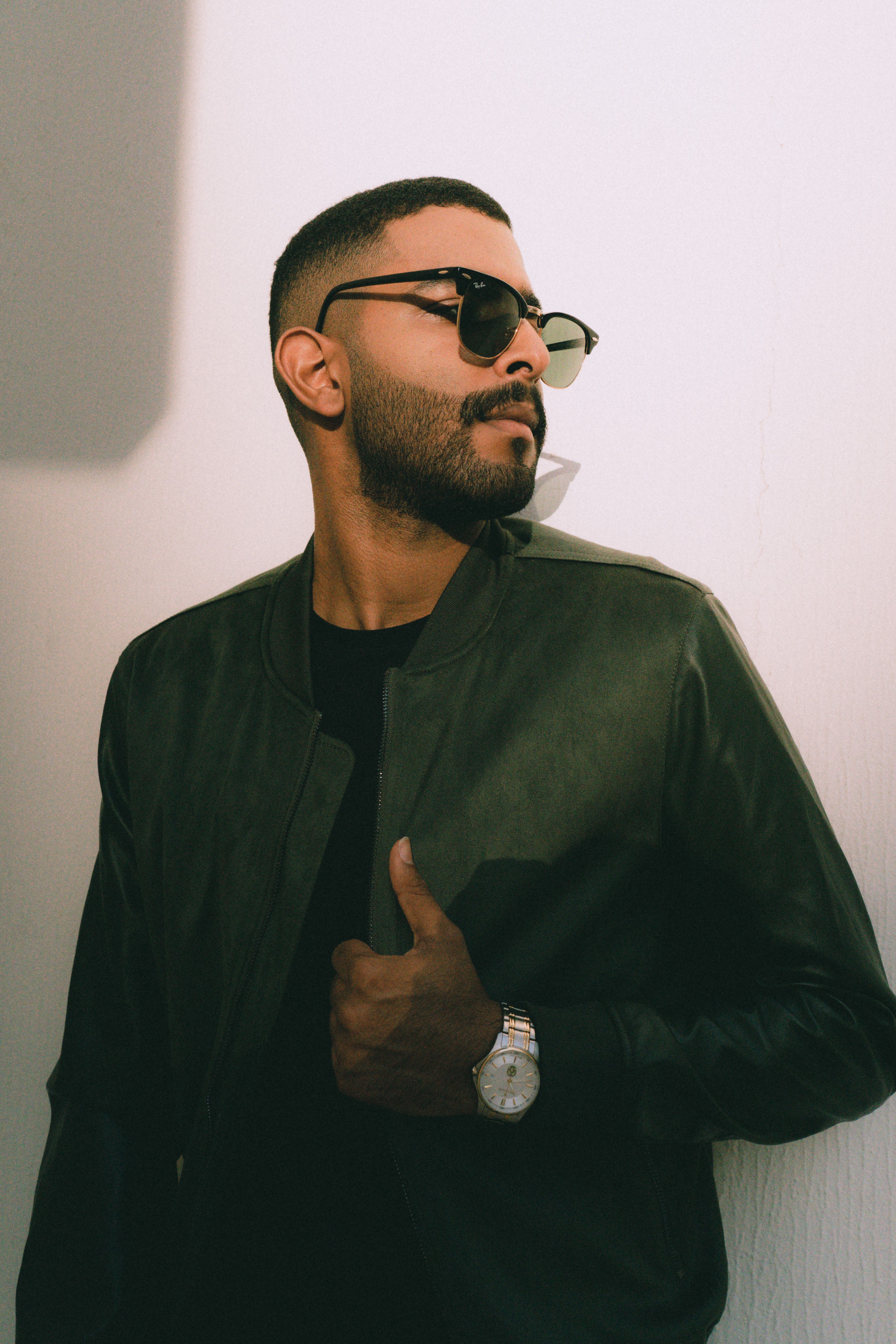 Fashionable man in sunglasses and leather jacket posing confidently against a sunlit wall.