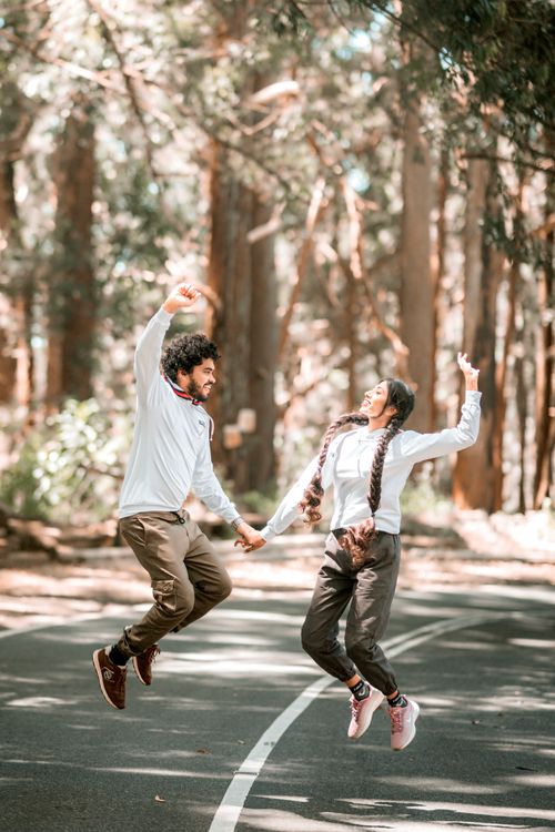 Free A couple joyfully jumps hand in hand on a serene forest road, surrounded by tall trees. Stock Photo