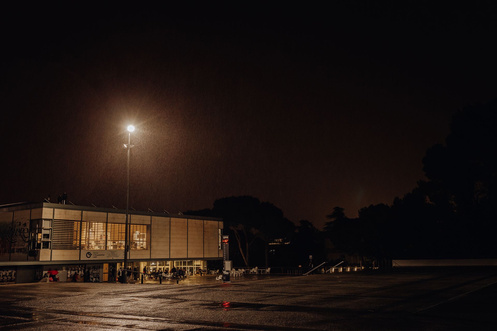 A nighttime urban scene featuring a modern building and wet street under street lights.