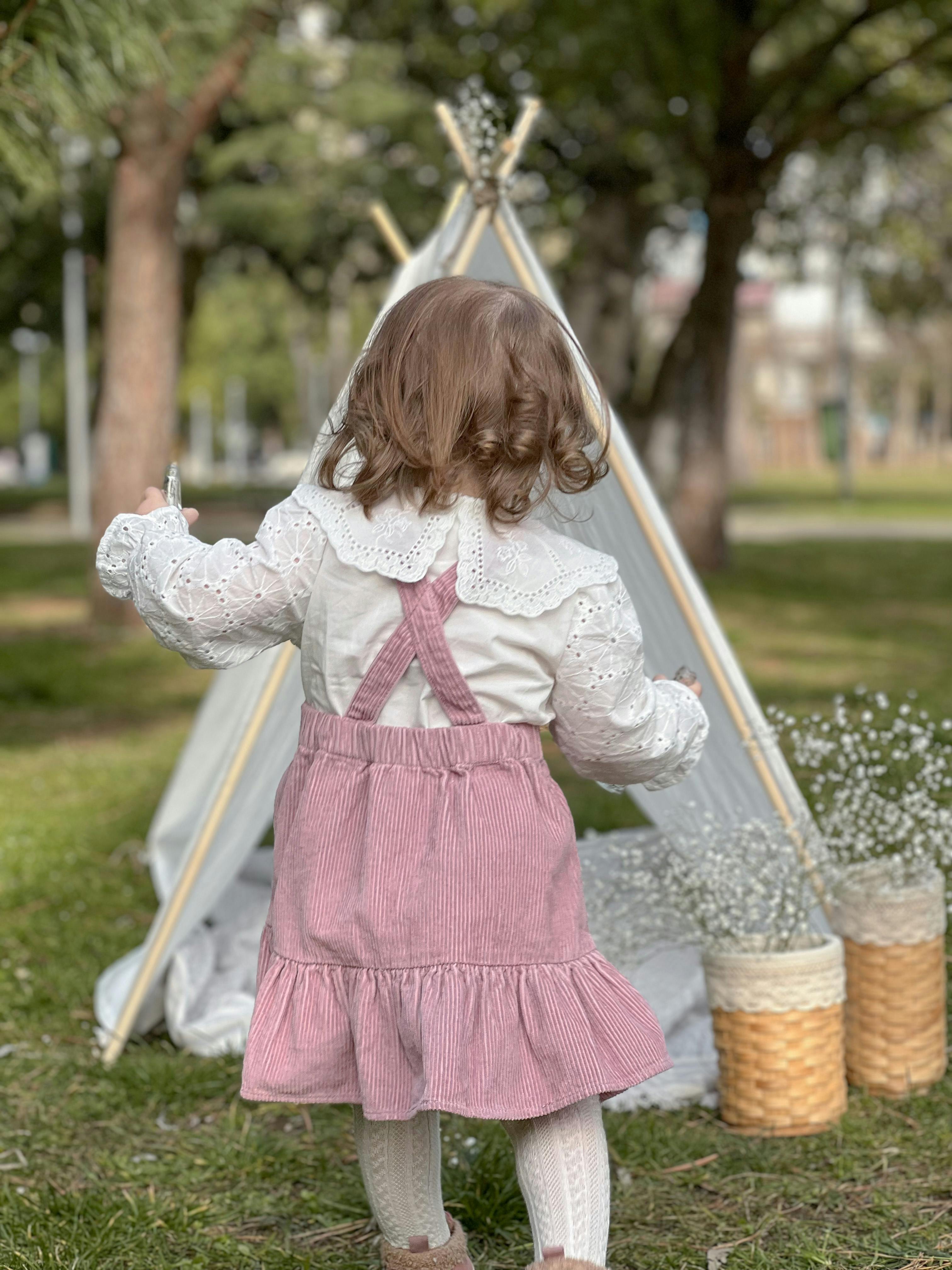 A young girl in a pink dress plays near a teepee tent in a park, capturing innocence and joy.