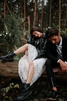 Stylish couple sitting on a log in the forest, combining elegance and casual attire.