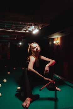 Moody portrait of a woman posing barefoot on a pool table under dim light.