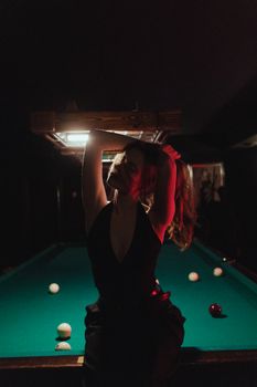 A stylish woman poses dramatically by a billiard table under a dim light, creating a moody nightlife atmosphere.