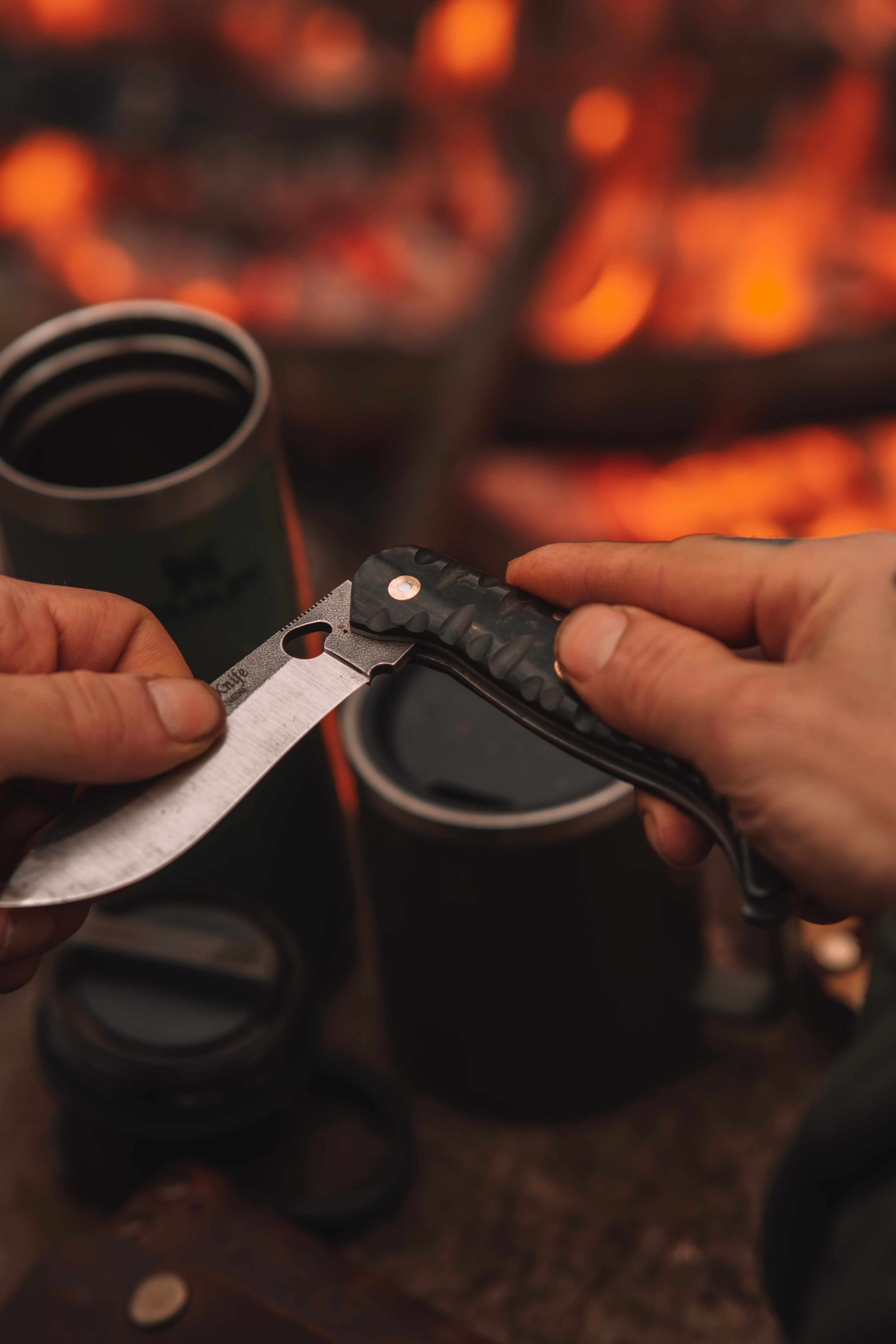 A person folding a knife with a camping scene in the background featuring a warm campfire.