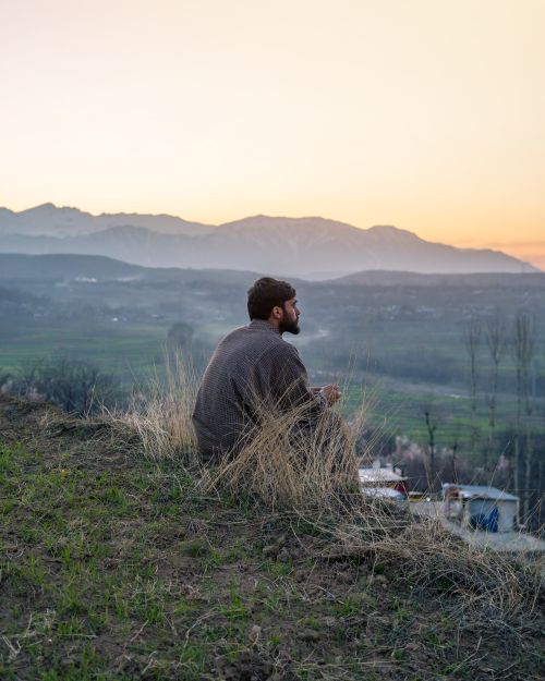Free A man sits on a grassy hill overlooking mountains at sunset, depicting tranquility in Srinagar. Stock Photo