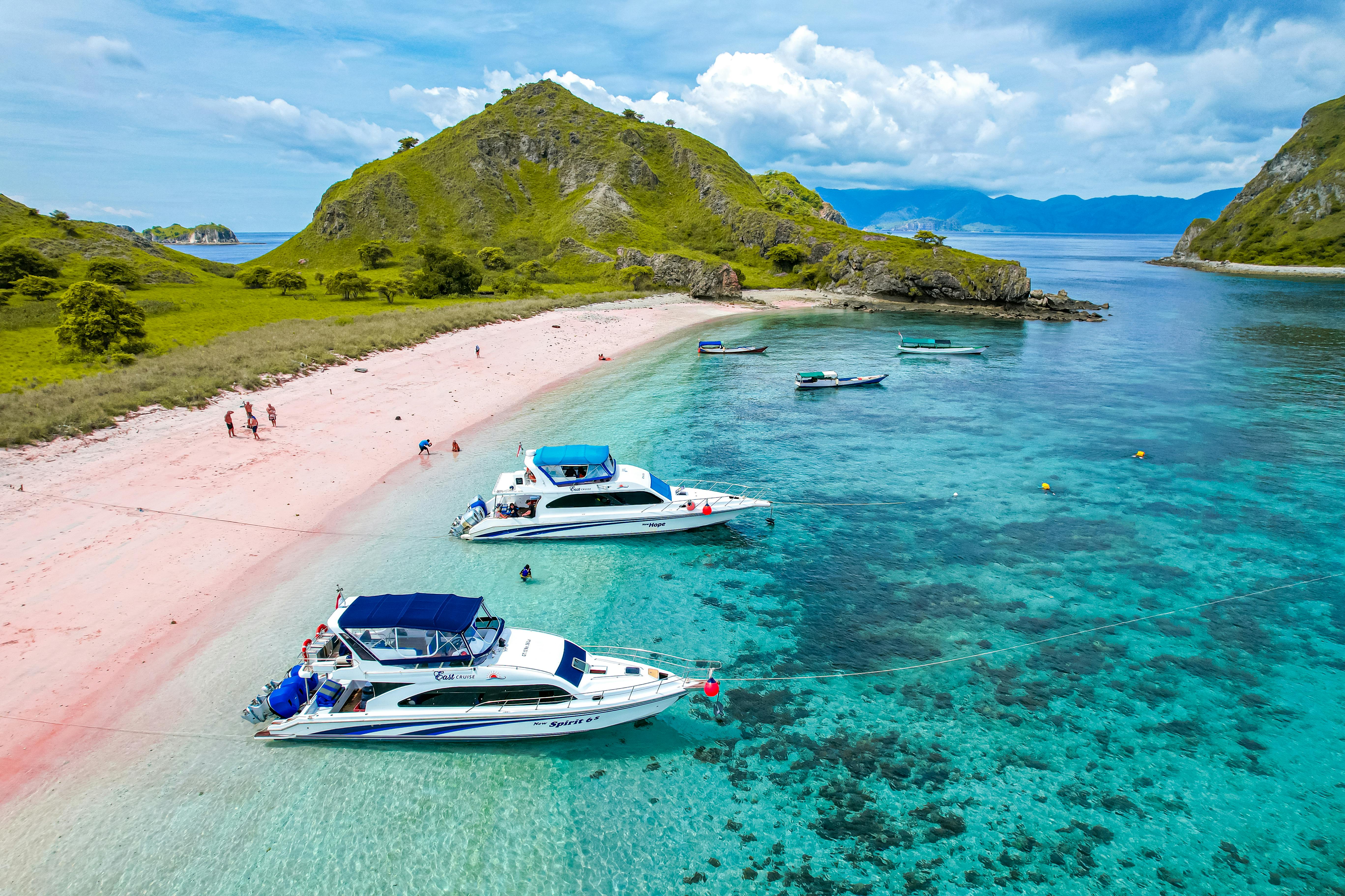 Meanwhile, Pink Beach in Labuan Bajo stretches along a pale pink shoreline with crystal-clear turquoise water, where several boats anchor offshore near a lush green hill under a bright blue sky.