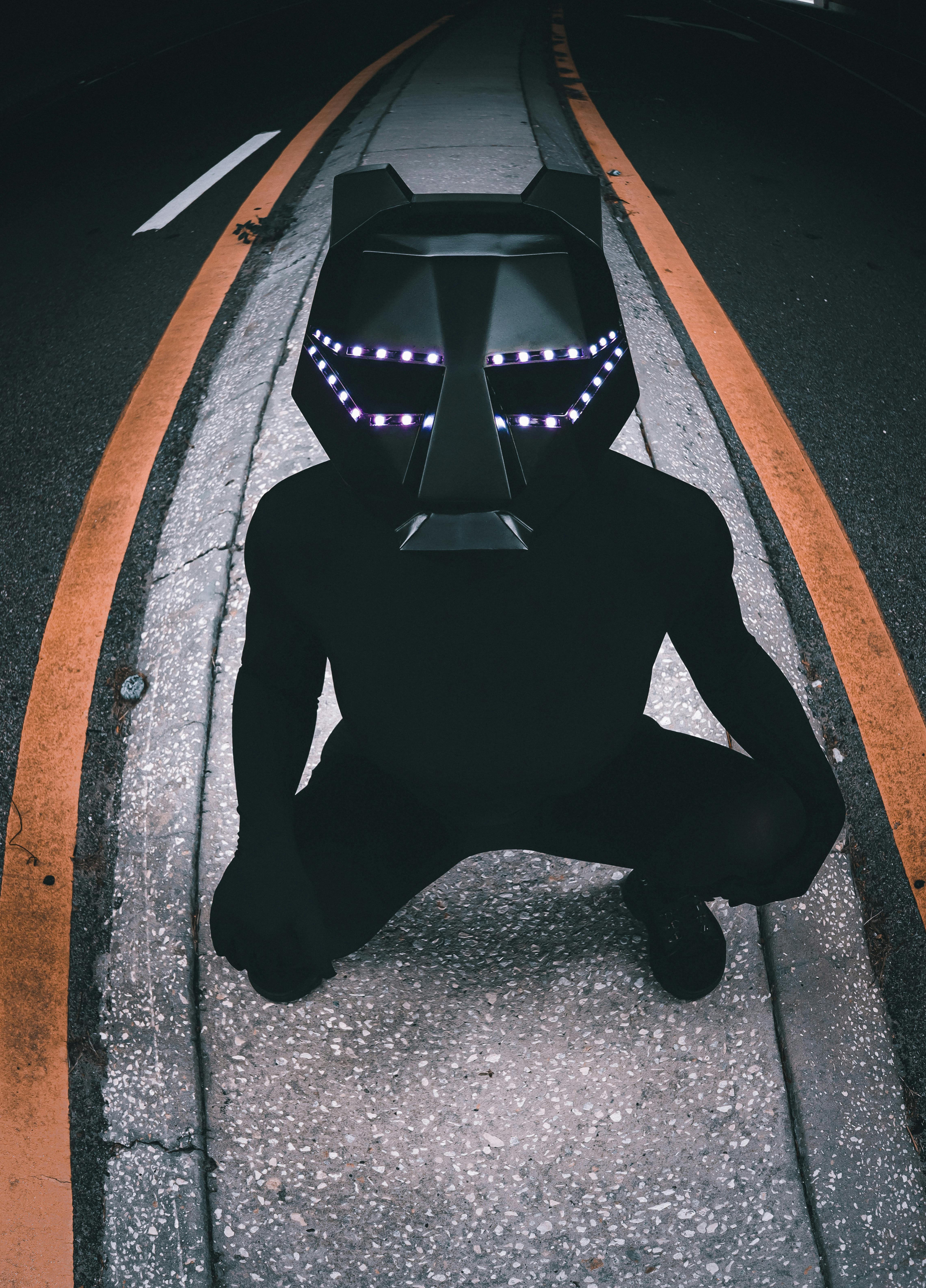 A person in a dark costume and LED helmet squatting on a city street with orange lines.