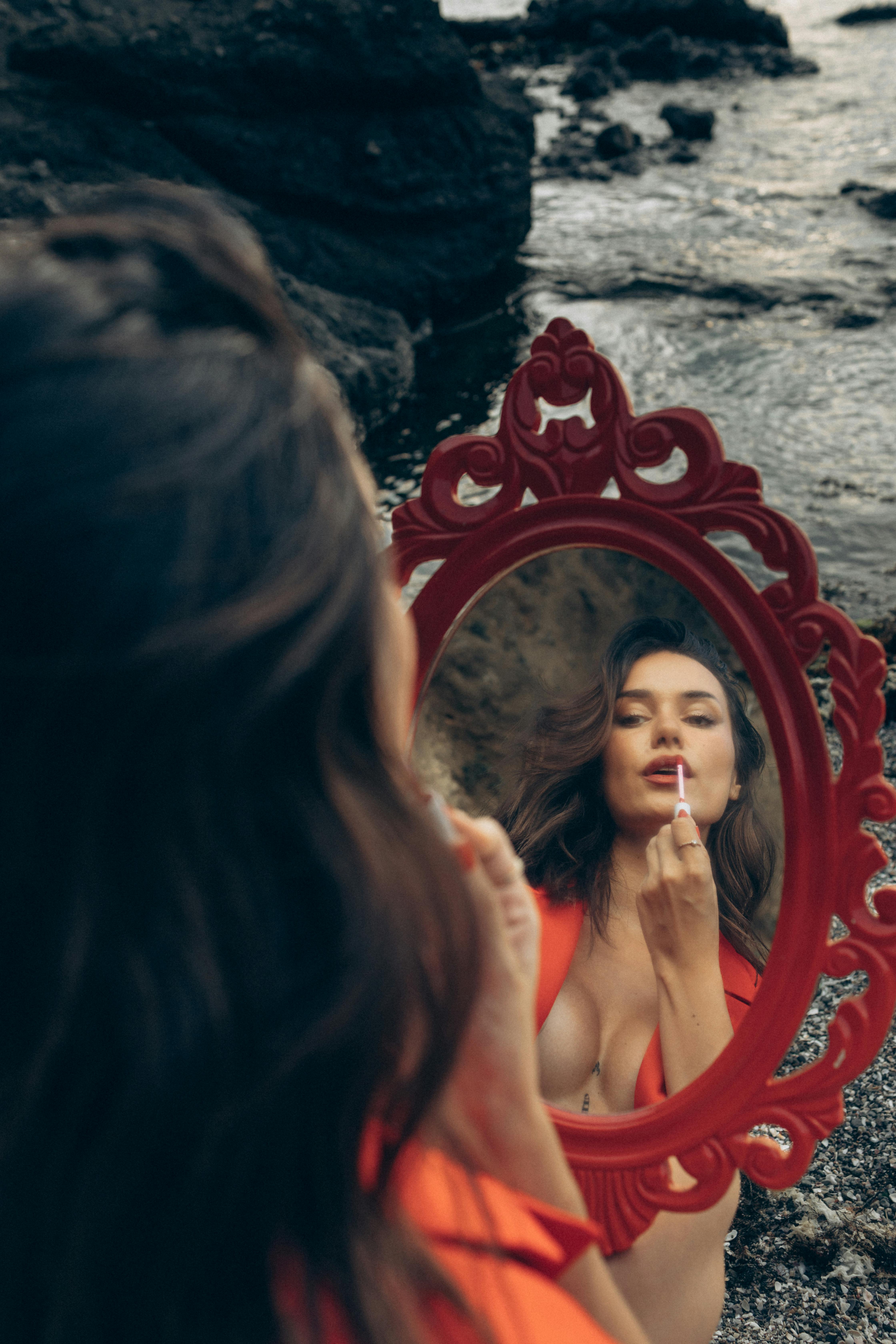 A woman applying lipstick, reflected in an ornate mirror by a rocky seashore.