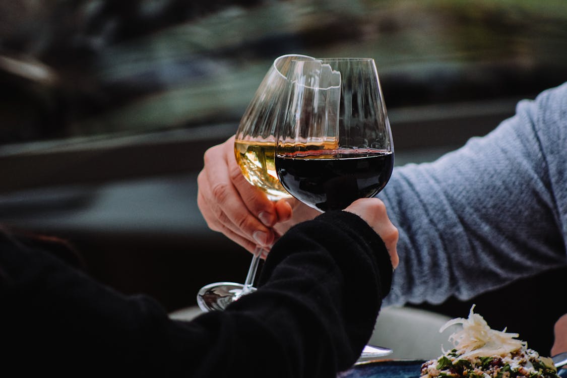 Free A couple clinking red and white wine glasses during an intimate outdoor moment.