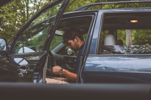 Free A man sits in a car with an open door in a wooded area, focusing attention ahead. Stock Photo