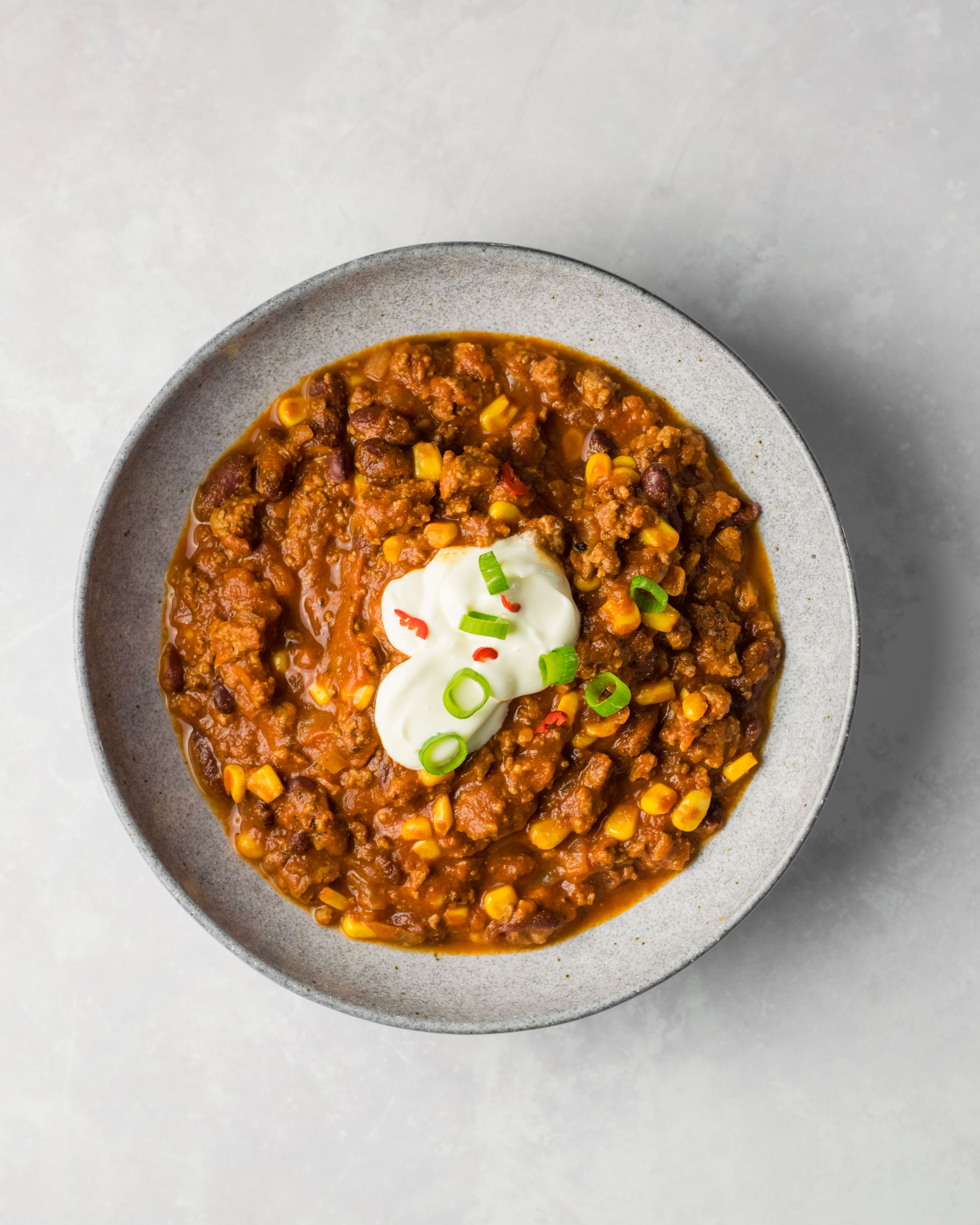 A bowl of red chili con carne with ground beef, beans, corn, sour cream, and sliced scallions