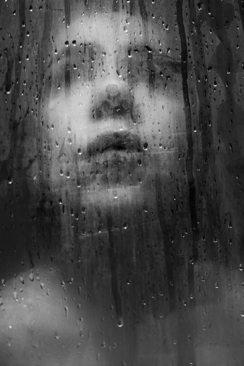 Free A moody black and white portrait of a woman viewed through a rain-covered window. Stock Photo